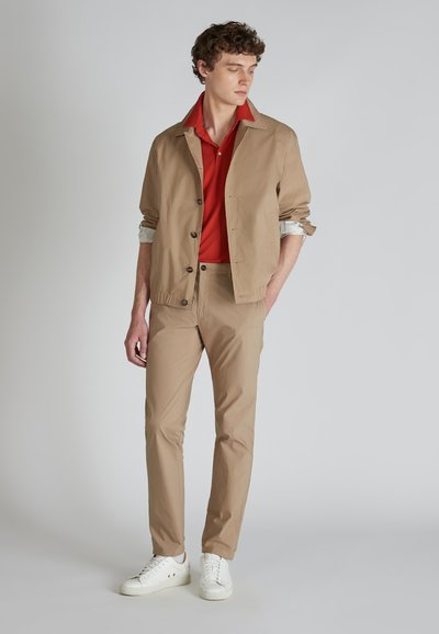 Chaqueta ligera beige con botones marrones, camiseta polo roja y pantalones beige. Zapatillas blancas completan el conjunto. Textura suave, diseño casual.
