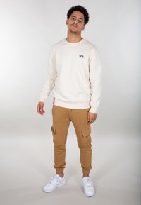 Lys beige sweatshirt med logo, parret med brune cargo bukser med lommer og snor. Hvide sneakers fuldender outfittet.