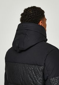 Giubbotto imbottito nero con cappuccio, texture trapuntata e motivo geometrico nella parte inferiore. Tessuto liscio con finitura opaca.