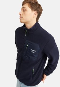 Chaqueta de forro polar azul marino con un cuello alto, bolsillo en el pecho con cremallera, acentos verdes en la cremallera y bolsillos laterales; textura suave y diseño casual.