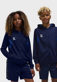 Marinet militær træningsdragter med tekstureret stof, udstyret med hættetrøjer med lynlås og kontrasterende blå accenter. Shorts fuldender det sporty look.