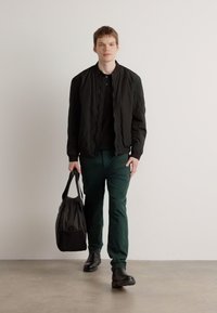 Chaqueta bomber acolchada negra sobre una camisa negra, combinada con pantalones verdes y zapatos negros. Sosteniendo una bolsa negra. Fondo neutro.