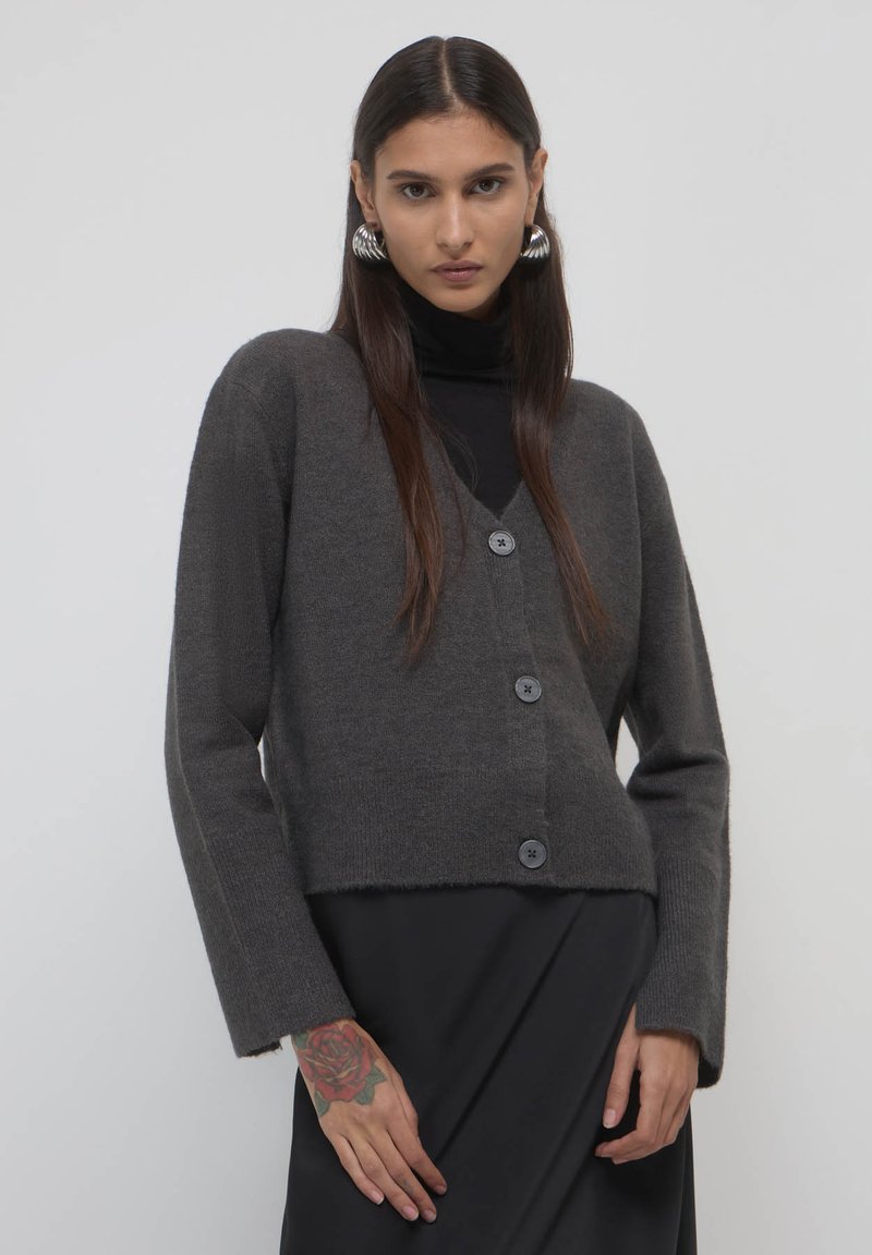 Dunkelgrauer Strickcardigan mit V-Ausschnitt, drei Knöpfen und langen Ärmeln, in cropped Design mit leicht ausgefranstem Saum.