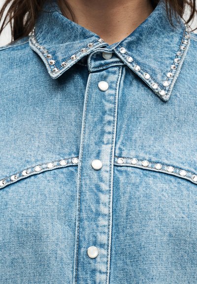 Ανοιχτό μπλε πουκάμισο τζιν με ασημένια τρουκς στον γιακά, λεπτομέρειες στο στήθος και λευκά κουμπιά με κούμπωμα μπροστά.