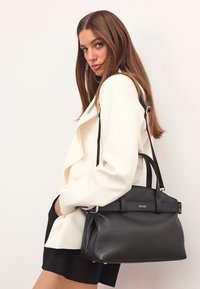 Fekete bőr kézitáska textúrált felülettel, arany hardver kiegészítőkkel, dupla fogantyúkkal és levehető vállpánttal. Fehér blézerral párosítható.