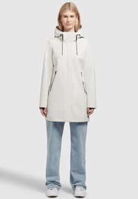 Parka impermeabile chiara con cappuccio, dotata di corde nere, due tasche laterali con zip e vestibilità dritta. Indossata sopra un paio di jeans casual.