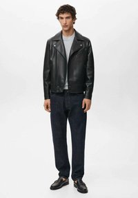 Veste de motard en cuir noir avec larges revers, poches zippées, portée sur un t-shirt gris, associée à un jean bleu foncé ample et des mocassins noirs.