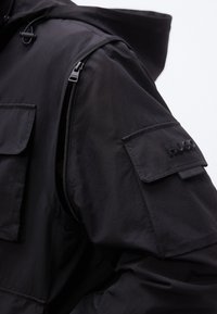 Μανίκι μαύρου μπουφάν με τσέπη με φερμουάρ και τσέπη με καπάκι που φέρει ανάγλυφο λογότυπο "HUGO" στο μπράτσο, σε λευκό φόντο.