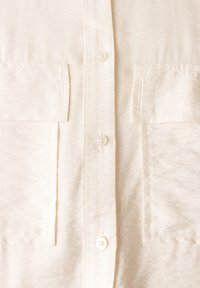 Camisa de botón color crema claro con dos bolsillos frontales, botones perlados y una tela texturada con un suave brillo.