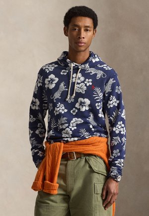 Homme portant un sweat à capuche bleu marine avec des imprimés floraux blancs et léopard, un pull orange noué autour de la taille, un pantalon cargo vert olive et une montre-bracelet.