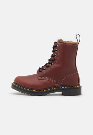 Bota al tobillo de cuero marrón con forro de piel sintética, cordones negros y costuras amarillas. Cuenta con suela con tacos y una lengüeta para tirar en la parte trasera.
