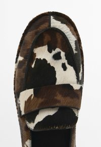 Moccasin-style shoe in brown and cream cowhide with a soft fur texture, featuring a rounded toe and a wide strap across the upper.