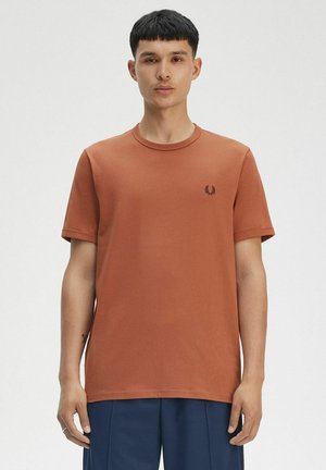 T-shirt en coton couleur rouille avec un col rond, des manches courtes, et un petit logo de marque noir sur la poitrine gauche. Design et texture simples.