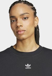 Černá mikina s kulatým výstřihem, ribbanou texturou a bílým vyšitým logem Adidas na hrudi. Hladká tkanina a standardní střih.