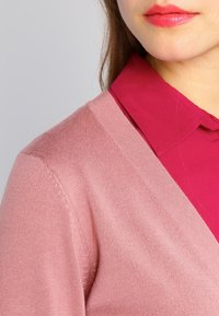 Un cardigan rose clair avec un design en V, fabriqué à partir d'un matériau en tricot doux, porté par-dessus une chemise col rose foncé. Texture lisse, couture subtile.