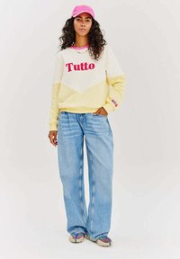 Krémová a žlutá mikina s grafikou "Tutto" v růžové, doplněná světle modrými širokými džíny a multikolorovými botami.
