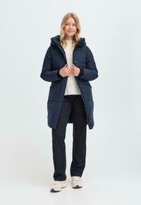 Mørkeblå puffjakke med hette, trykknapplukking, sidelommer og quiltet tekstur, iført over en lys genser og sorte bukser.
