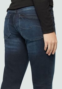 Dunkelblaue Denim-Jeans mit schmaler Passform, die zwei Gesäßtaschen und dezente Nähdetails aufweisen, aus einem seitlichen Blickwinkel aufgenommen.