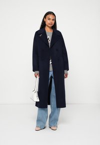 Woman wearing a long navy double-breasted coat, gray sweater, wide-leg jeans, holding a white handbag, standing against a white background.