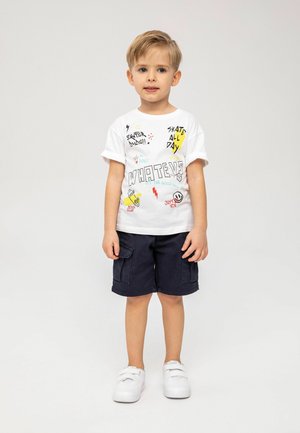 Biała bawełniana koszulka z wieloma kolorowymi grafikami, połączona z granatowymi szortami cargo i białymi sneakerami na rzep.
