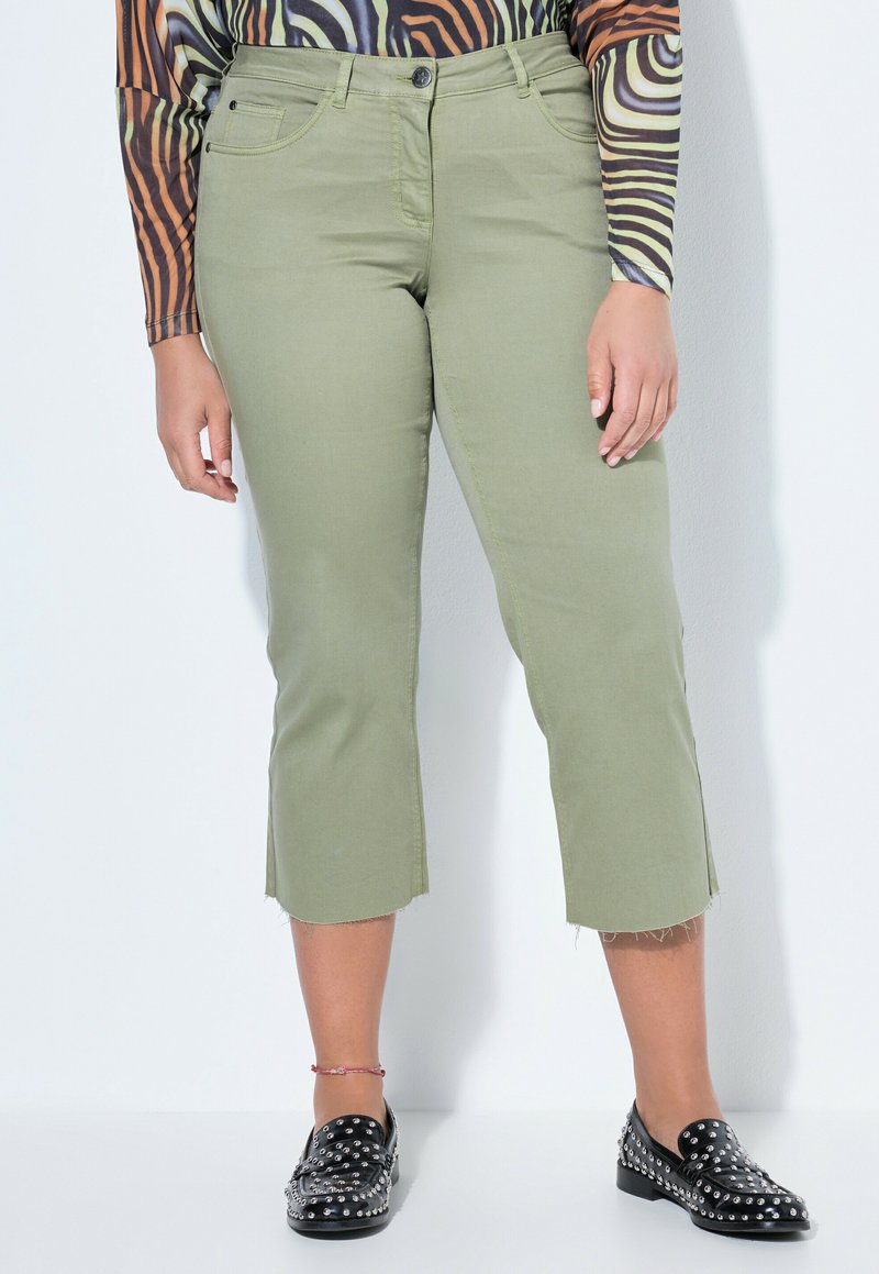 Person trägt olivgrüne, gekürzte Hosen mit ausgefransten Säumen, schwarze Loafers mit Nieten und ein langärmliges Zebramuster-Oberteil, steht vor einer hellen Wand.