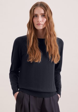 Jonge vrouw met lang golvend rood haar, gekleed in een zwarte trui met lange mouwen en donkere geplooide broek, staand met handen in de zakken.