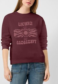 Sweat-shirt bordeaux avec un design graphique incluant les mots "VIVRE LE MOMENT" et un motif de soleil circulaire. Poignets et ourlet côtelés.