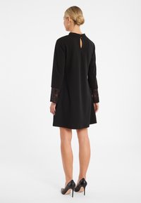 Schwarzes Kleid mit langen Ärmeln, Spitzenakzenten an den Manschetten und einem Schlüsselloch-Detail im Rücken. Kombiniert mit schwarzen High Heels.