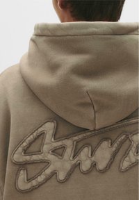 Beige hoodie med et tekstureret broderet logo på ryggen, med en rummelig hætte og blødt, glat stof.