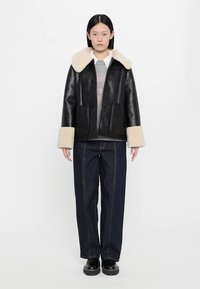 Giacca di pelle nera con colletto e polsini in pelliccia ecologica crema, tasche frontali, indossata sopra un maglione grigio e abbinata a pantaloni di jeans scuri e stivali neri.