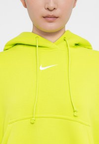 Nike kapuce spilgti neona zaļā krāsā, ar priekšējo kengūra kabatu, auklītēm un mazāku balto logo uz krūšu. Mīksta tekstūra.