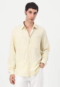 Cămașă butonată de culoare galben deschis, cu un model texturat, cu dungi și o buzunare pe piept. Mâneci lungi și tiv rotunjit. Purată cu pantaloni albi.