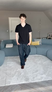 Čierne tričko s výstrihom do V, modré voľné nohavice a hnedé topánky. Pozadie obsahuje zakrivenú modrú pohovku a textúrový biely koberec.