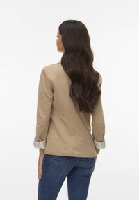 Blazer beige su misura con maniche lunghe, caratterizzato da polsini arrotolati a righe. Indossato sopra un paio di jeans in denim blu, mostrato da dietro.