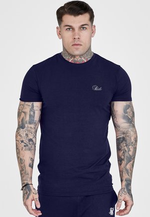 Mann med tatoveringer på hals og armer iført en tettsittende mørkeblå t-skjorte med korte ermer og matchende mørkeblå bukser på en ensfarget bakgrunn.