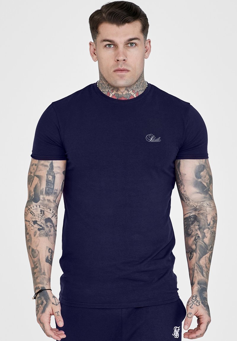 Homme avec des tatouages sur le cou et les bras portant un t-shirt bleu marine ajusté à manches courtes et un pantalon bleu marine assorti sur un fond uni.