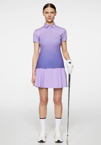 Lilla gradient golf polo med knaplukning og korte ærmer, parret med en matchende plisseret nederdel. Hvide golfhandsker og sko.