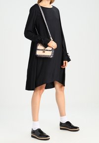 Robe noire à manches longues avec une coupe ample, présentant des poignets froncés. Associée à un petit sac à main crème et des chaussures noires en vernis avec une semelle en bois.