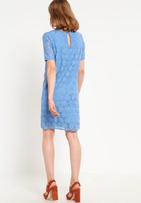 Short blue lace dress with short sleeves, featuring a keyhole back detail and a straight cut. Worn with brown block-heeled sandals.