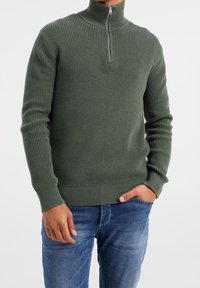 Maglione a coste verde con collo alto e chiusura con zip, dotato di maniche lunghe e una vita aderente, abbinato a jeans blu.