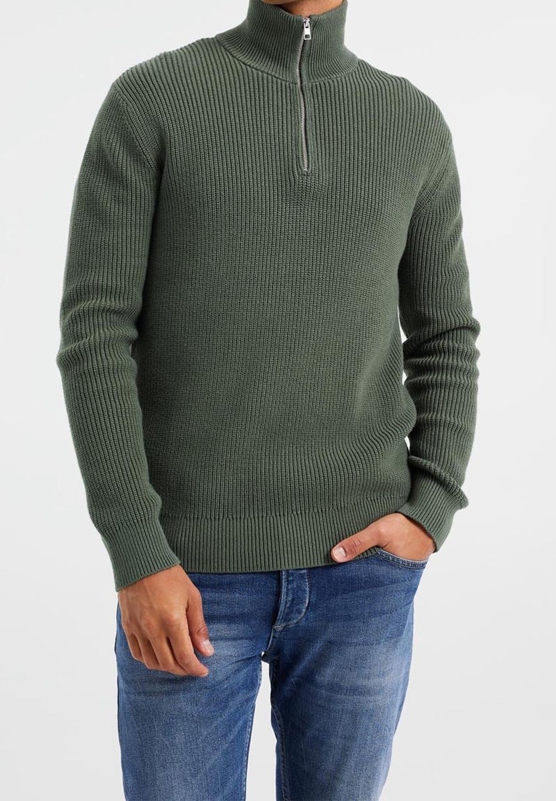 Maglione a coste verde con collo alto e chiusura con zip, dotato di maniche lunghe e una vita aderente, abbinato a jeans blu.