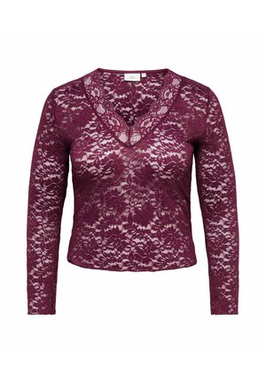 Vrouw met krullend donker haar draagt een paarse kanten top met lange mouwen en een zwarte leren midirok met een voorsplit, gecombineerd met paarse schoenen.