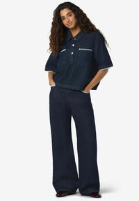 Temno denim obleka s kratkimi rokavi in ovratnikom, z dvema žepoma spredaj in širokimi hlačami v istem slogu. Robovi z obrobo dodajo teksturo.