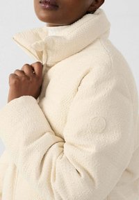 Chaqueta acolchada crema con superficie texturizada, cuello alto y cierre de cremallera. Presenta un logo sutil en la manga. Tejido suave.