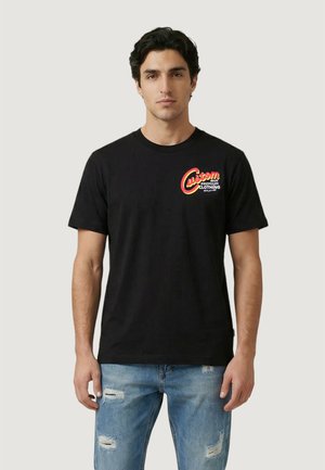 Uomo in piedi che indossa una maglietta nera con il logo "Custom Premium Clothing" rosso e giallo e jeans blu strappati su sfondo chiaro.