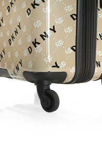 Maleta rígida en color beige con un patrón de logotipo DKNY en blanco y negro. Cuenta con dos ruedas negras y un cierre de cremallera texturizado.