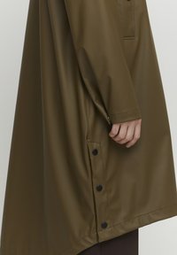 Trench coat de culoare verde măsliniu, impermeabil, cu o croială lejeră. Include un buzunar lateral, nasturi la tiv și o textură netedă, lucioasă.