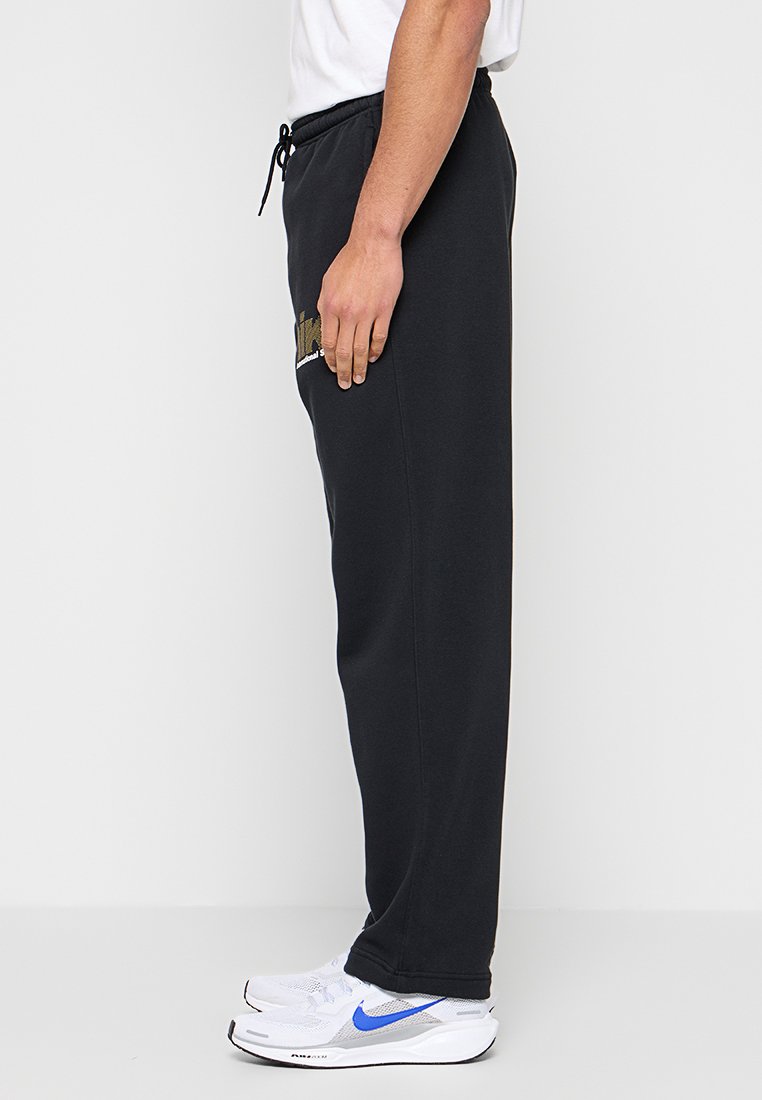 Personne debout portant un pantalon de survêtement noir avec une taille à cordon et des baskets blanches avec des accents bleus, sur un fond blanc uni.