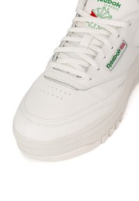 Baltas odinis sportbačius su tekstūruota paviršiaus, suapvalintu nosimi, plokščiais raišteliais ir žaliu Reebok logotipu su raudonomis detalėmis ant liežuvėlio.