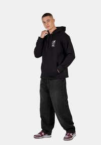 Sudadera negra con bolsillo frontal, logo blanco en el pecho, combinada con jeans negros holgados y zapatillas rojas y grises. Tejido suave y corte relajado.